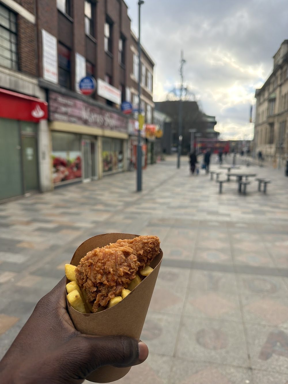 A paper cone of fried chicken and chips held on the high street outside